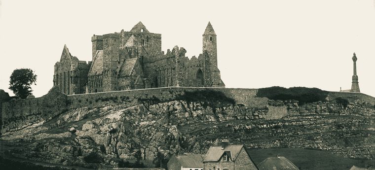 The Rock of Cashel photographed about 1900: the cathedral and round tower on their limestone outcrop, with thatched and slated cottages and stone-walled fields in the foreground.